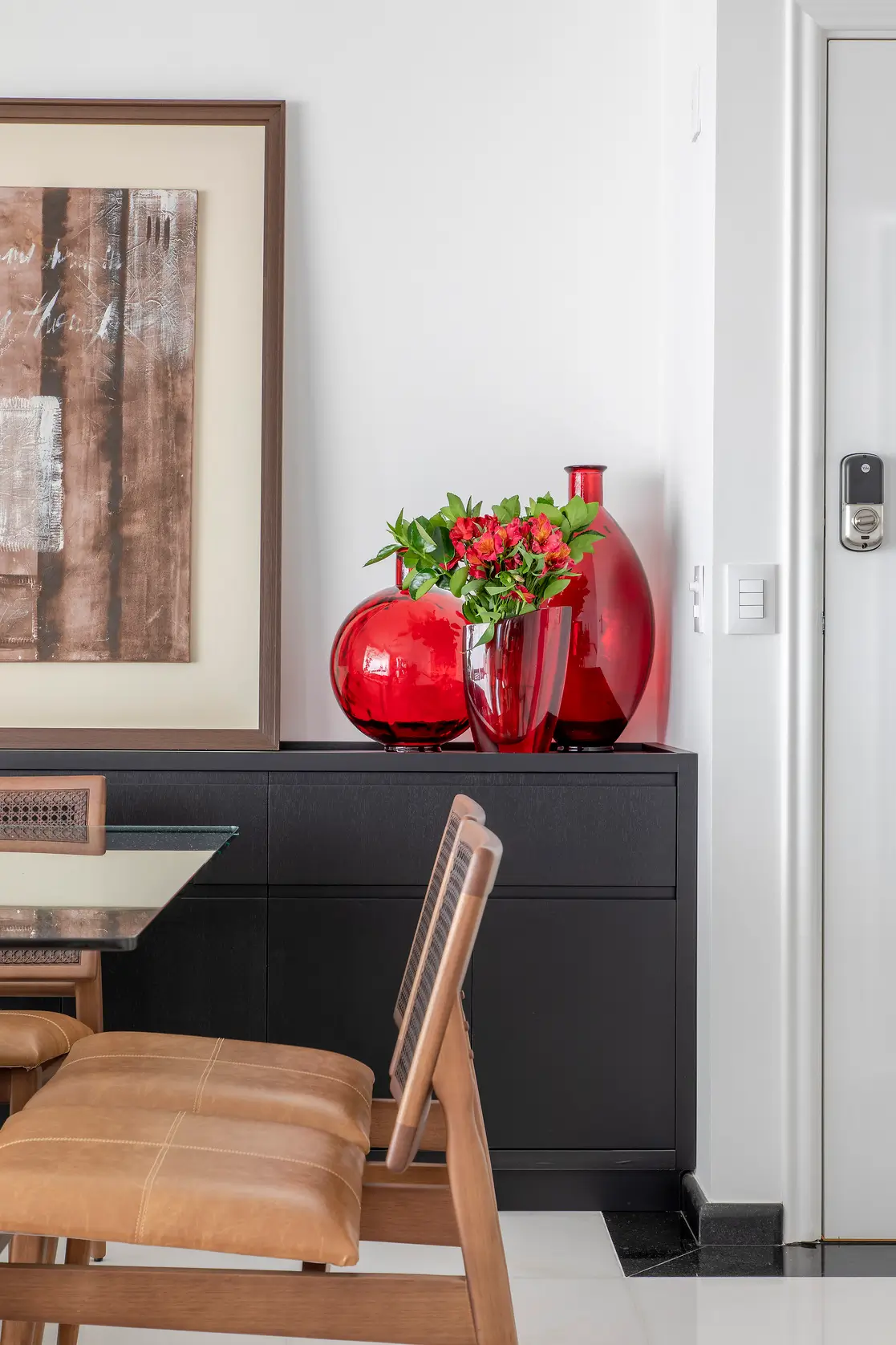 Aparador preto com vasos de vidro vermelho e flores ao lado da mesa de jantar em projeto da MIS Arquitetura